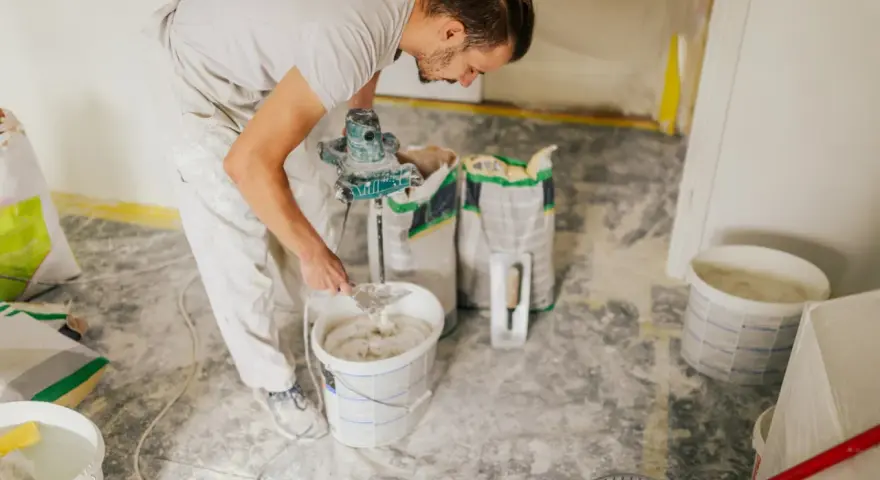 Un ouvrier prépare de l'enduit béton cellulaire dans un seau avec un mélangeur. Des sacs de poudre sont à proximité.