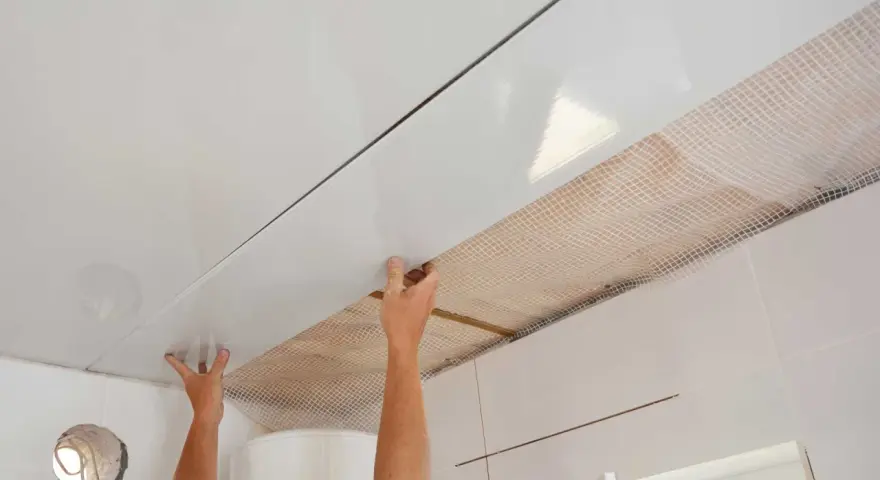 Pose de lambris PVC au plafond avec clips. Les mains ajustent une lame blanche brillante sur une structure, révélant un treillis blanc en dessous.