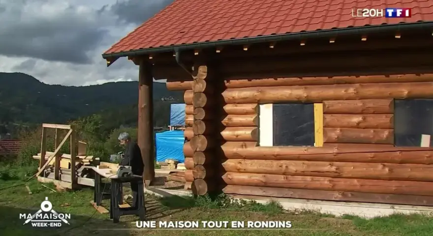 Une maison en rondins de bois en construction, avec un homme travaillant à proximité. Le toit est rouge et le paysage est montagneux.