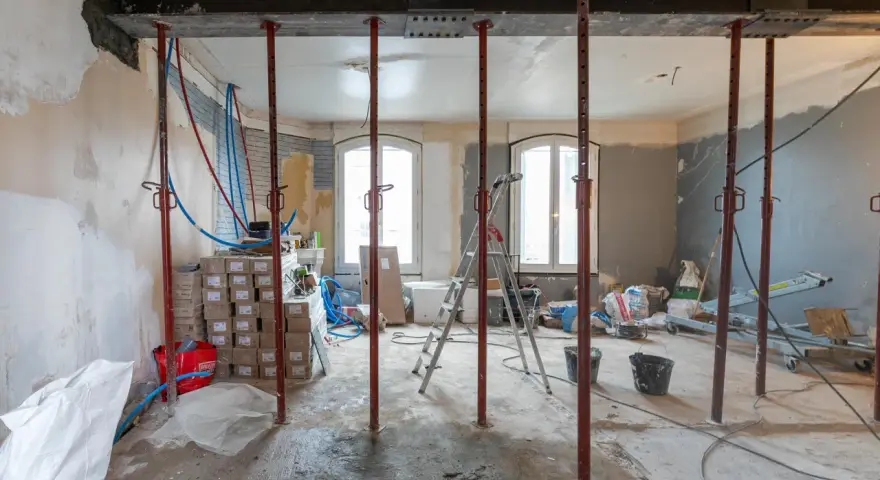 Chantier d'une maison phenix 1970 : poutres métalliques, étais, tuyaux et matériaux de construction dans une pièce en rénovation.