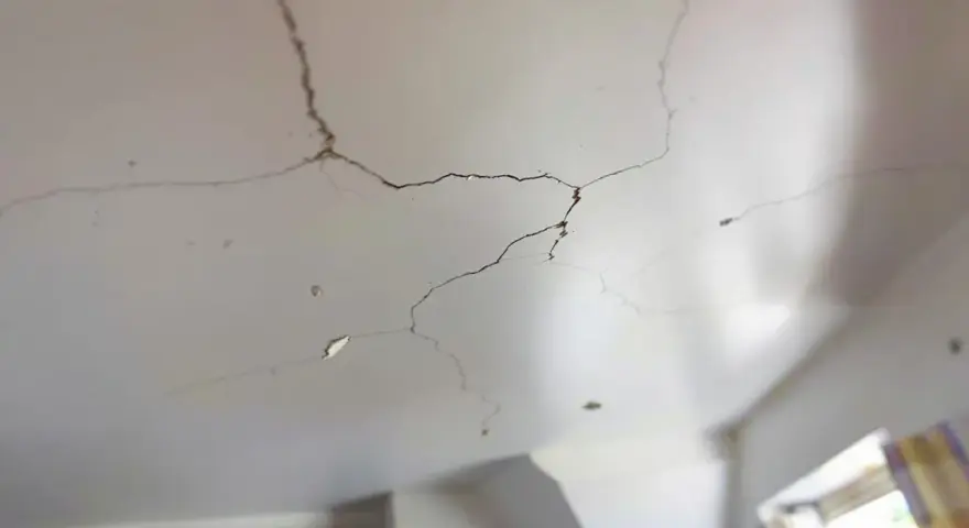 Un plafond qui s'effrite, révélant des fissures sombres et des morceaux de plâtre tombés. La lumière douce éclaire les imperfections.