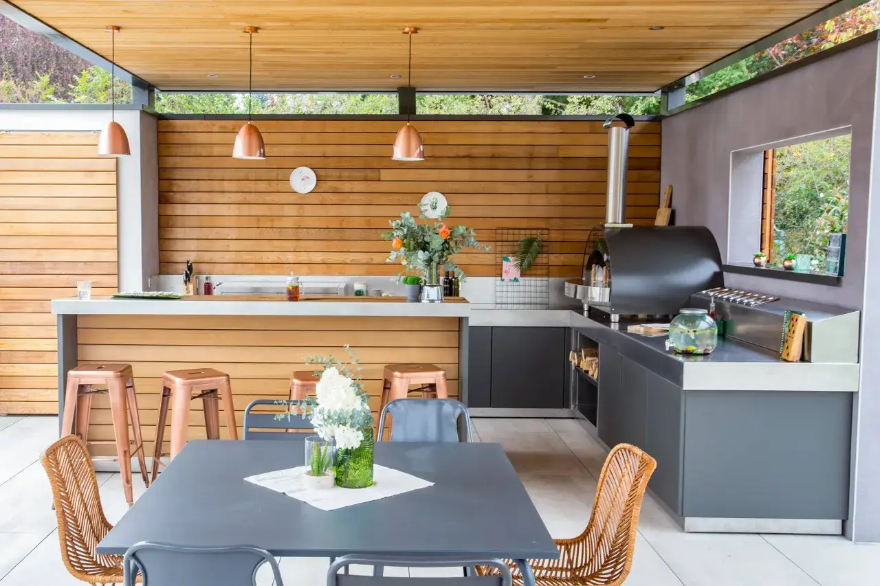 Un espace repas extérieur moderne avec un four à pizza, un bar et une table. Ce plan cuisine d'été est parfait pour recevoir.