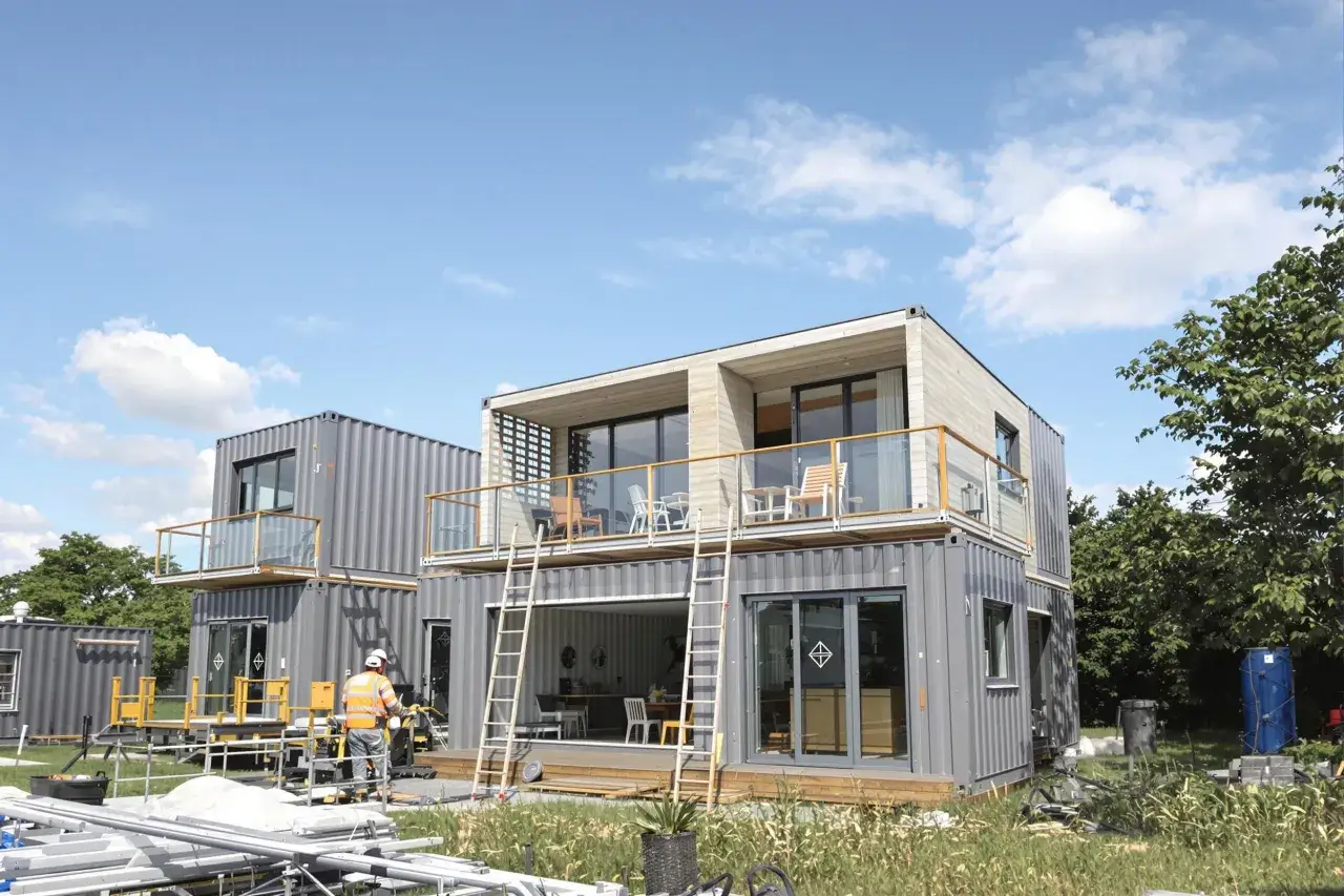 Maison en container prix : construction moderne à deux étages avec balcons, fenêtres panoramiques et un ouvrier en gilet orange.
