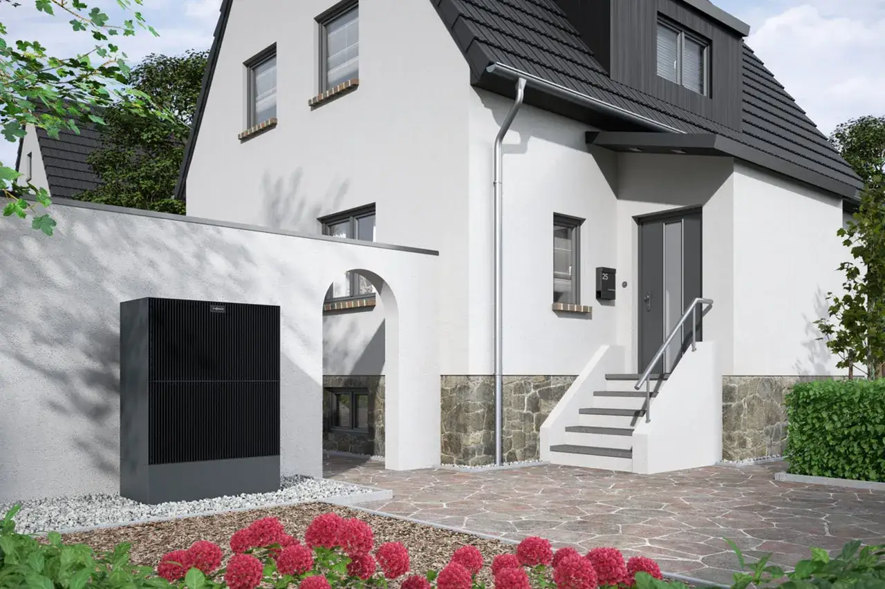Une maison moderne avec une pompe à chaleur monobloc noire installée à côté d'un mur blanc. Des fleurs roses décorent le premier plan.