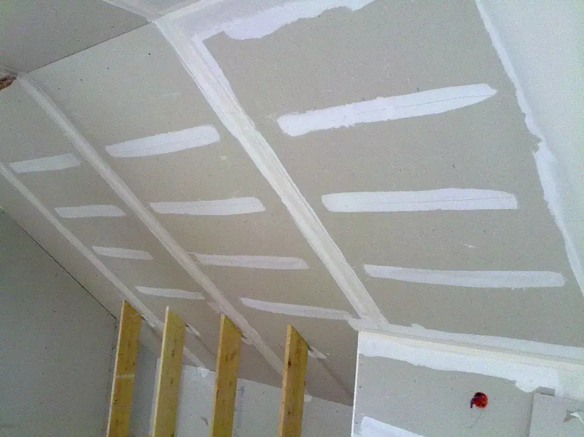 Plafond en cours de finition avec bande de placo et enduit blanc. Des tasseaux en bois soutiennent la structure.