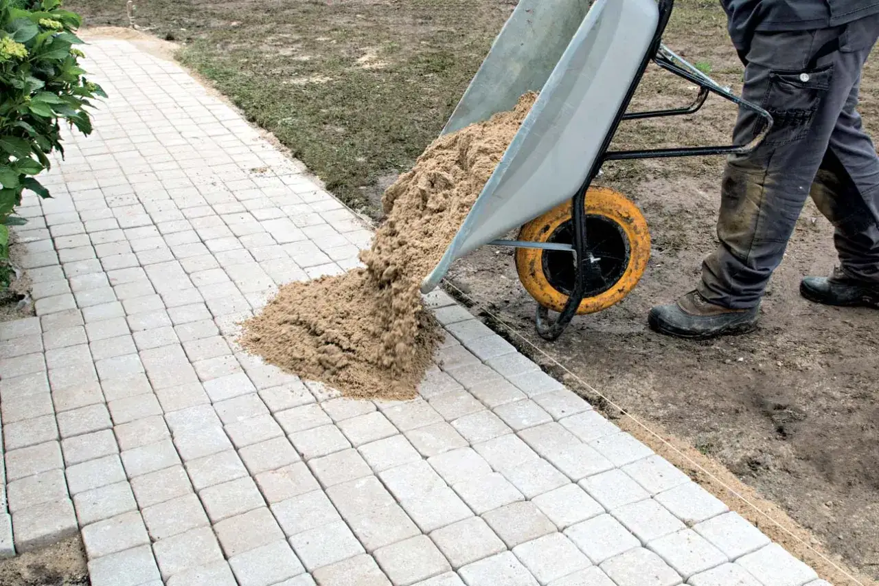Pose de pavé extérieur : une brouette déverse du sable sur un chemin en cours de construction, prêt pour le pavage.