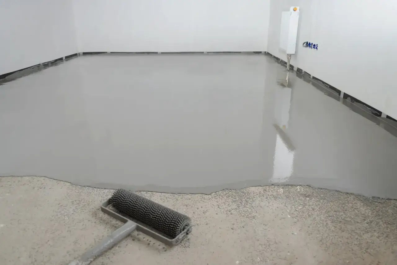 Un sol lisse et gris fraîchement coulé, résultat d'un ragréage autonivelant. Un rouleau à picots est posé sur le béton brut, prêt pour la suite des travaux.