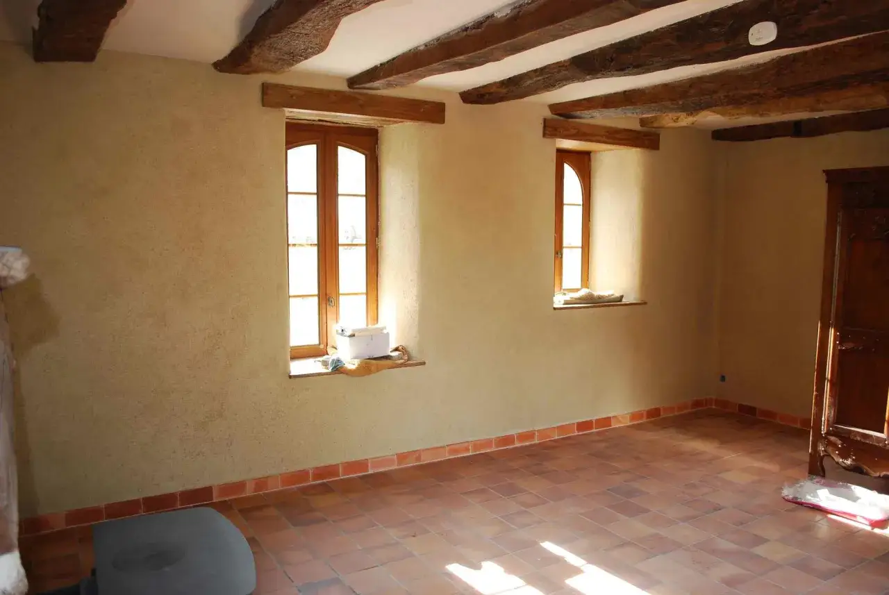 L'isolation d'un mur intérieur dans une maison ancienne, avec poutres apparentes, deux fenêtres et un sol en terre cuite.