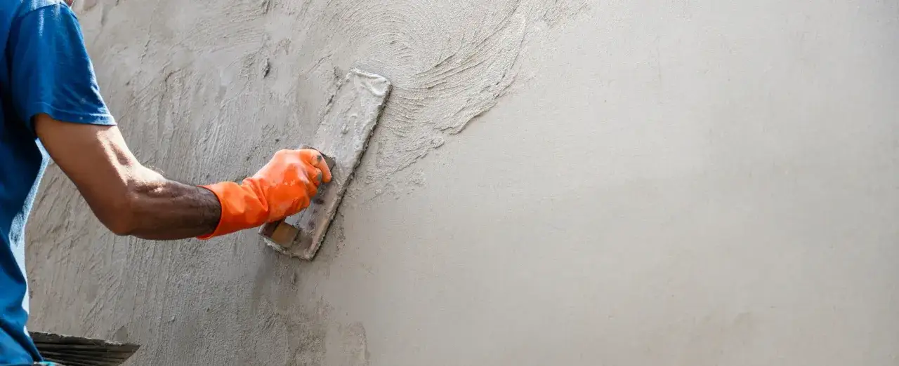 Main tenant une taloche, un ouvrier applique l'enduit sur un mur en parpaing. L'épaisseur de l'enduit est visible.
