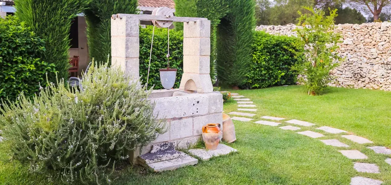 Un vieux puits en pierre avec un seau suspendu, entouré de verdure luxuriante et d'un chemin de dalles.