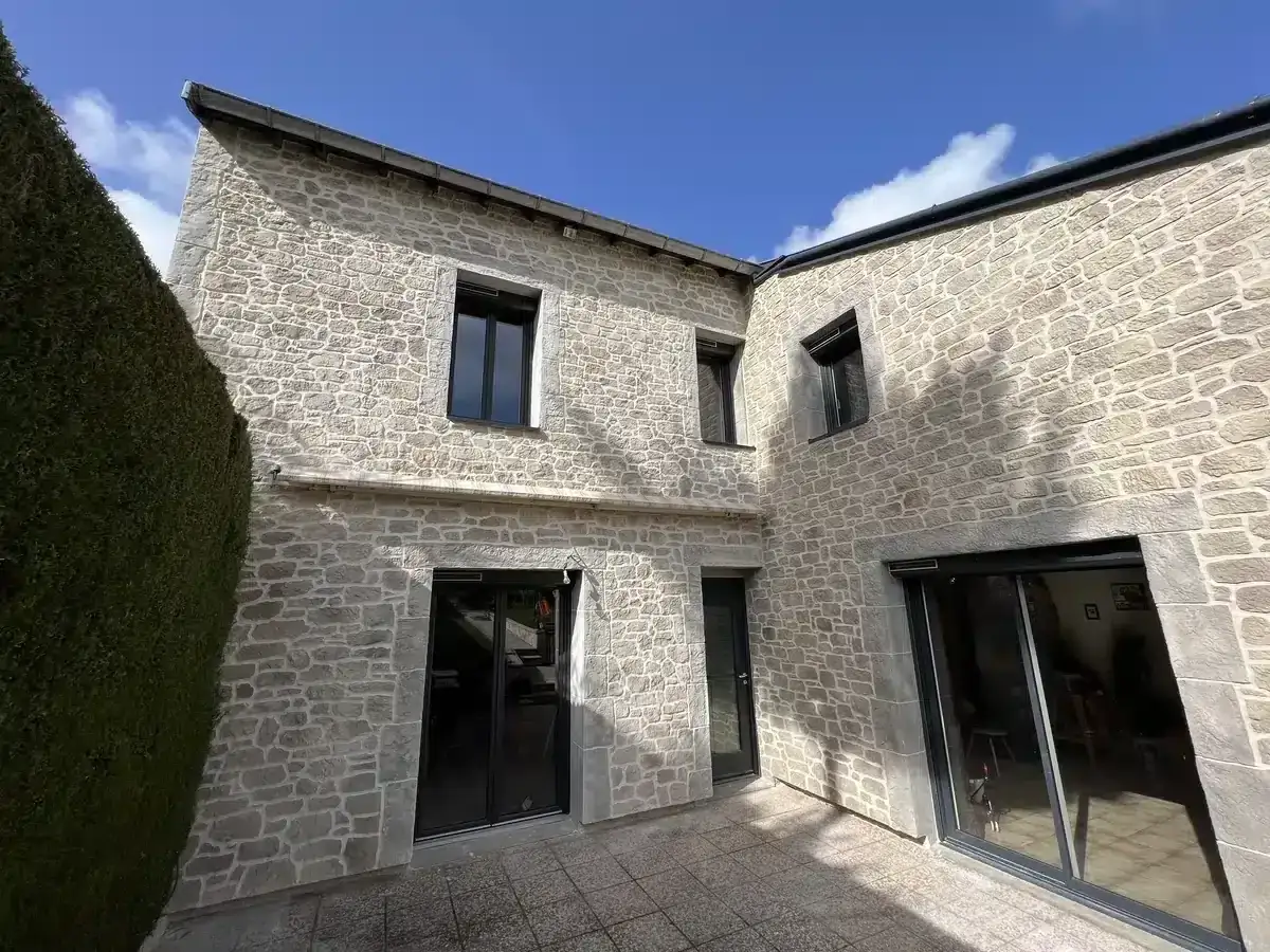 Maison en pierre avec un mur extérieur irrégulier, une haie verte luxuriante et un ciel bleu clair.
