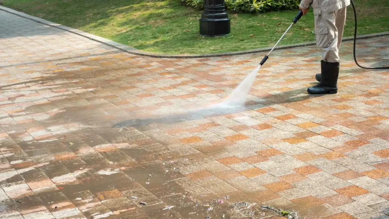 Un homme en bottes nettoie une terrasse en béton avec un nettoyeur haute pression. L'eau projetée révèle la propreté retrouvée.