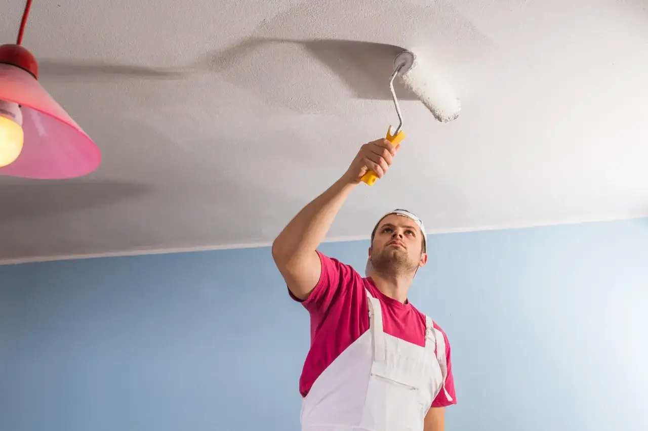 Un homme en salopette blanche et t-shirt rose peint le plafond avec un rouleau. Il réfléchit à quelle peinture pour plafond choisir pour un résultat parfait.