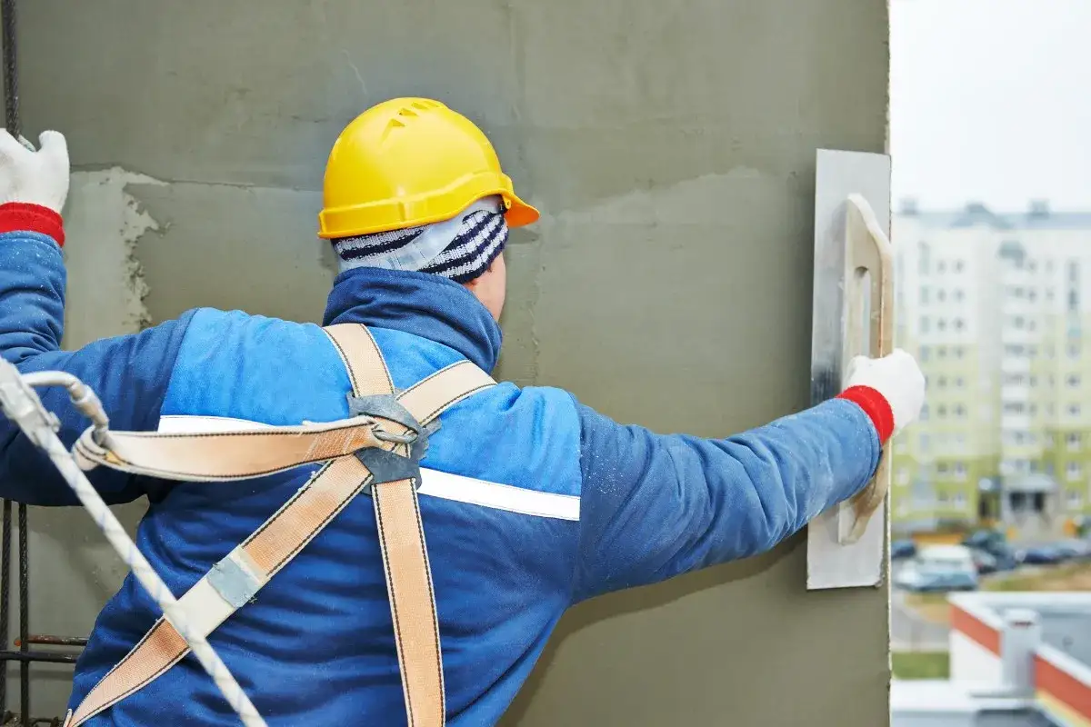 Ouvrier en b&acirc;timent appliquant un enduit sur un mur ext&eacute;rieur, assurant l'isolation sous enduit. Il porte un casque jaune et un harnais de s&eacute;curit&eacute;.