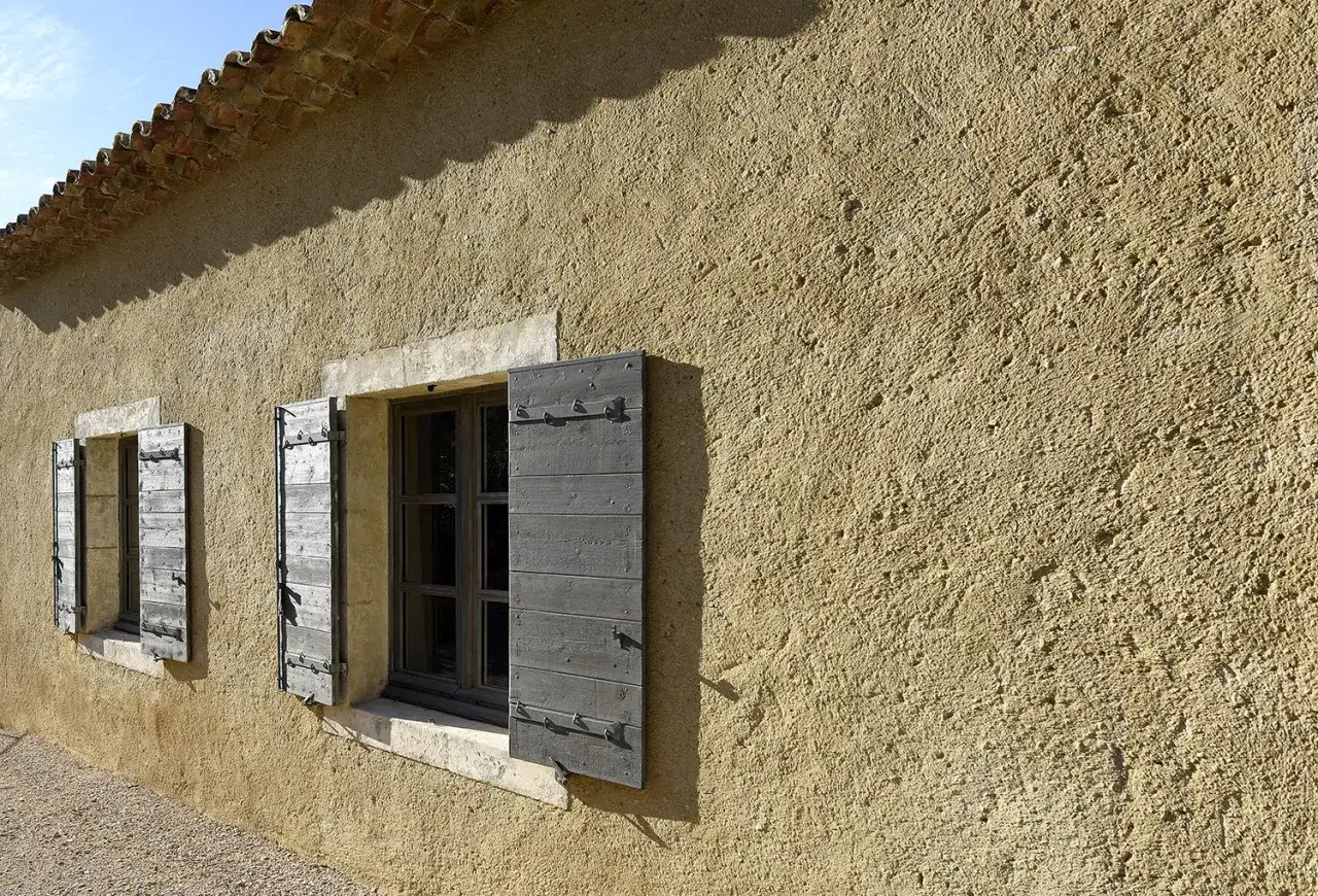Façade d'une maison avec un enduit à la chaux extérieur texturé, deux fenêtres aux volets en bois gris ouverts, et un toit de tuiles romanes.