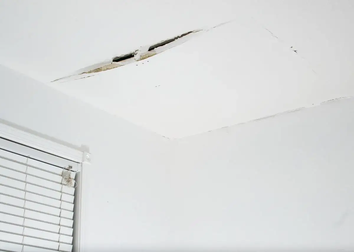 Fissure béante dans un plafond en placo, nécessitant une réparation. Un coin de fenêtre avec stores est visible.