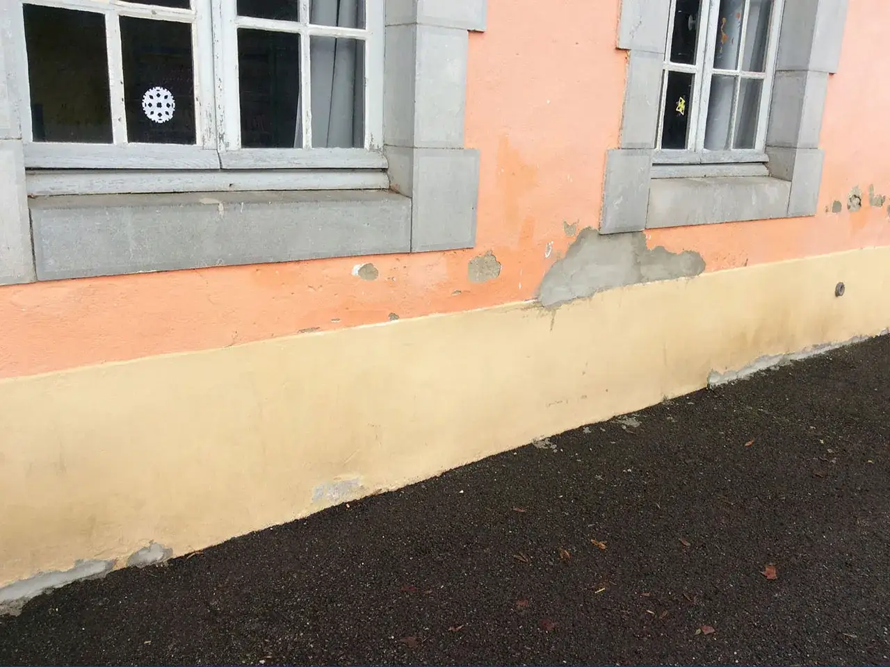 Détérioration du crépi due à la remontée capillaire du sol. Le bas du mur est décoloré et écaillé, près du sol sombre.