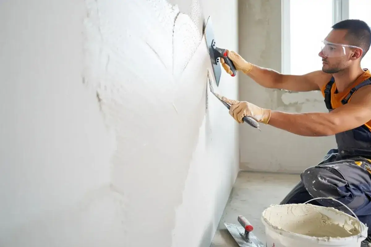 Un ouvrier applique un enduit sur un mur humide avec une truelle et une taloche. Le travail progresse.