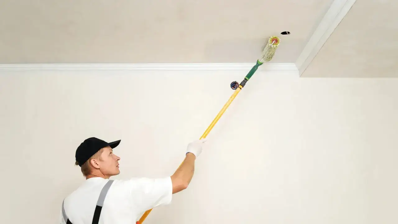 Un peintre utilise un rouleau à long manche pour peindre un plafond. Il porte une casquette noire et des bretelles.