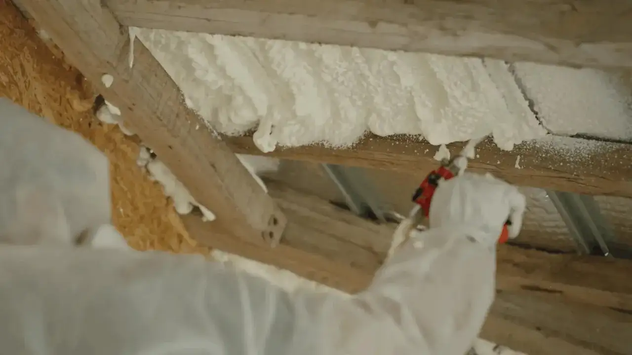 Application d'isolation mousse projet&eacute;e sur des poutres en bois. Le travailleur porte une combinaison de protection.