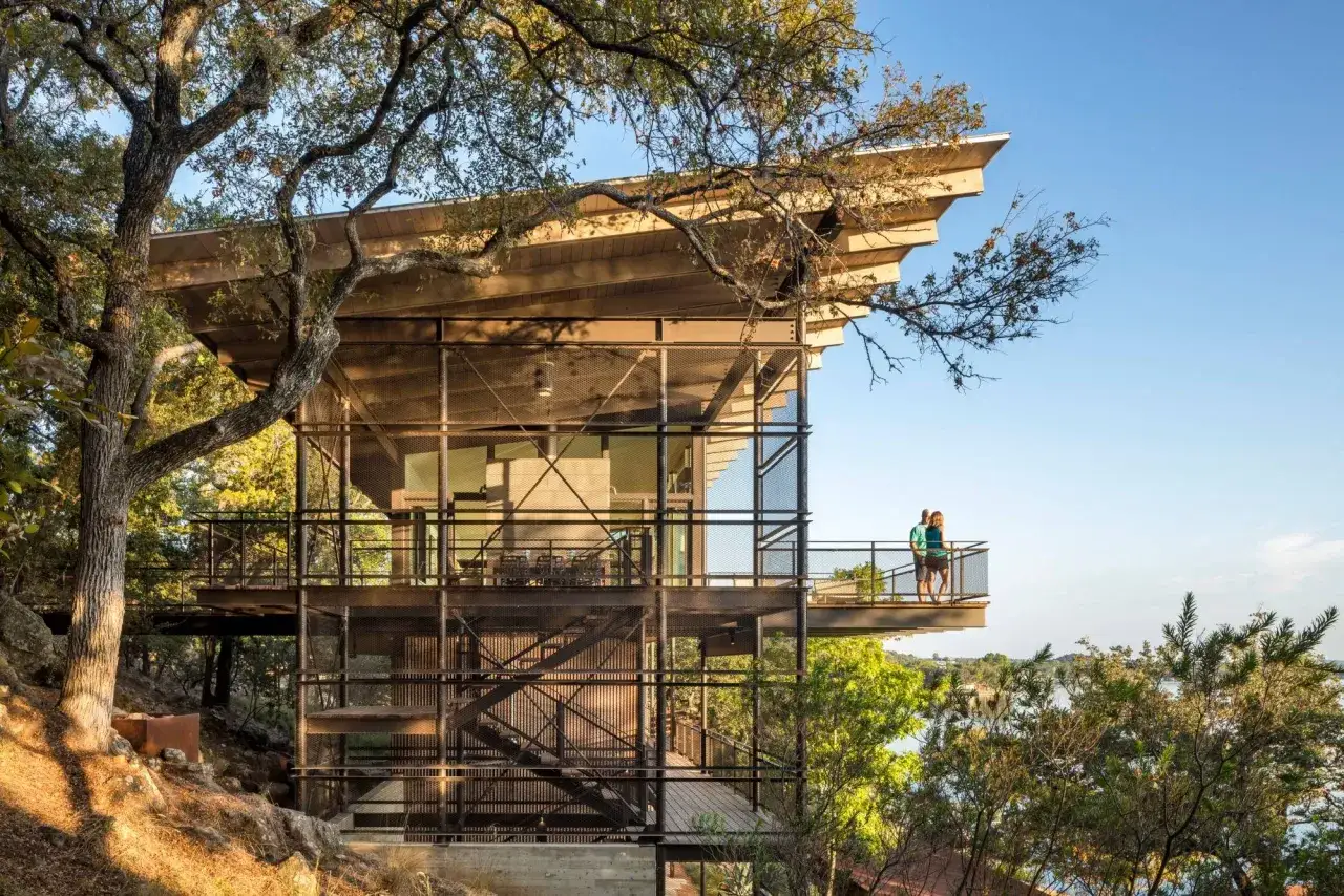 Une maison sur terrain en pente raide, avec une structure métallique et des balcons offrant une vue sur un lac. Deux personnes admirent le paysage.