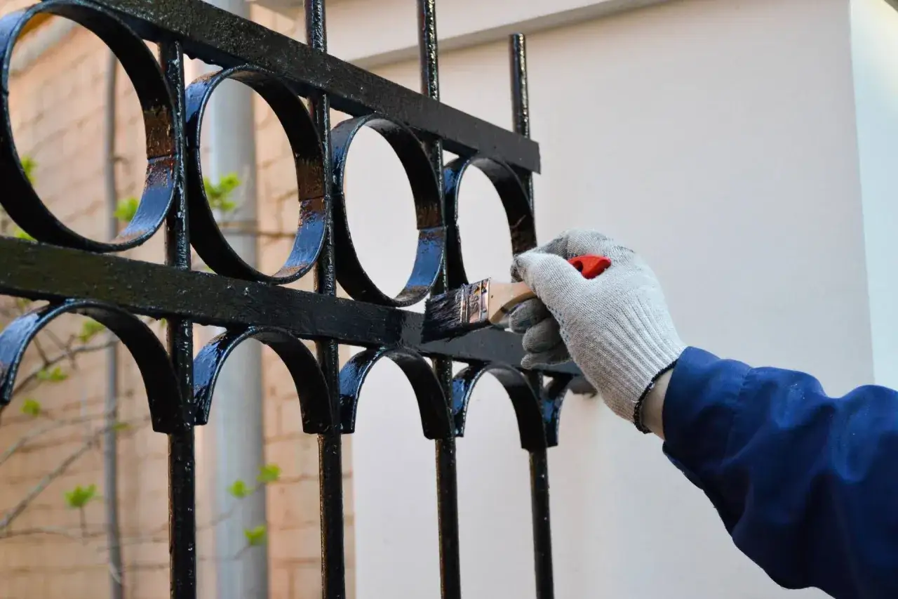 Main tenant, ganté, applique une couche de peinture noire sur une grille en fer forgé ornée de cercles. Le nettoyage du fer forgé avant peinture est essentiel pour un résultat durable.