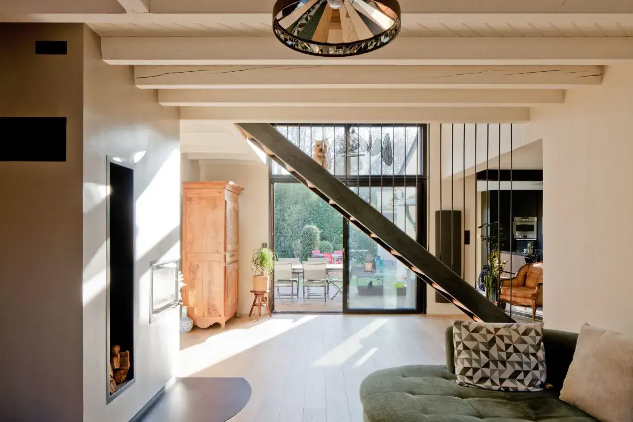 Intérieur lumineux d'une maison aux caractéristiques des années 70, avec poutres apparentes, cheminée moderne, et un chien curieux regardant par la fenêtre.