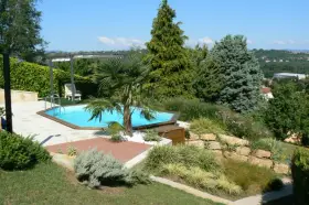 Piscine aux formes douces sur un terrain en pente, entourée de végétation luxuriante et d'une terrasse en bois.
