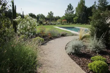 Une allée de jardin originale serpente vers une piscine, bordée de lavande, de fleurs blanches et d'arbustes verts.