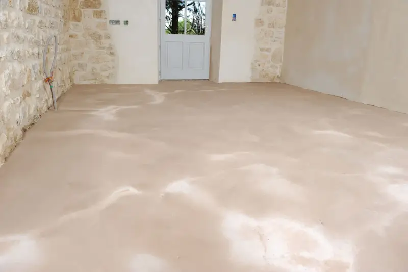 Sol fraîchement ragréé, l'épaisseur du ragréage intérieur est visible sur le sol brut. Les murs en pierre et la porte blanche annoncent une rénovation.